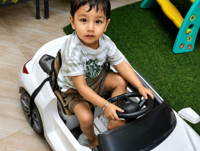 Little boy sitting at the wheel of a white toy car on artificial grass