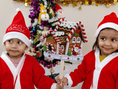 Two children in Santa outfits holding a gingerbread house by the Christmas tree
